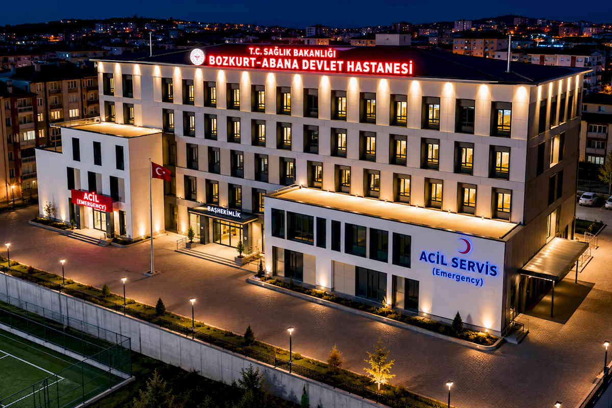 Kastamonu Bozkurt-Abana Devlet Hastanesi'nin gece saatlerinde drone ile çekilmiş genel görünümü. Fotoğrafta aydınlatılmış hastane binası, acil servis, başhekimlik girişi, çevre düzenlemesi ve otopark alanı görülmektedir.